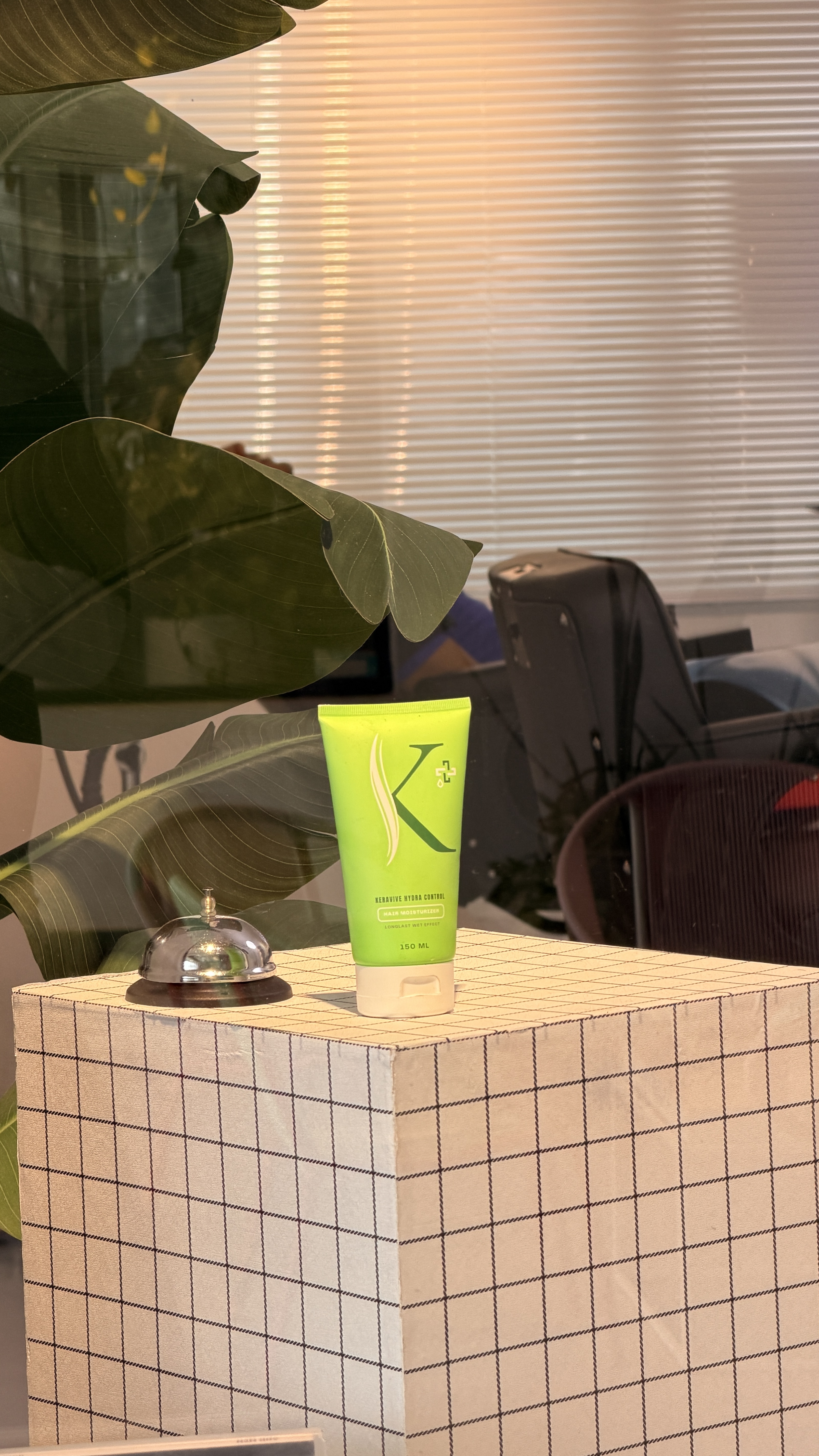 A grooming product and a service bell on a tiled counter, beside green leaves and a barber chair